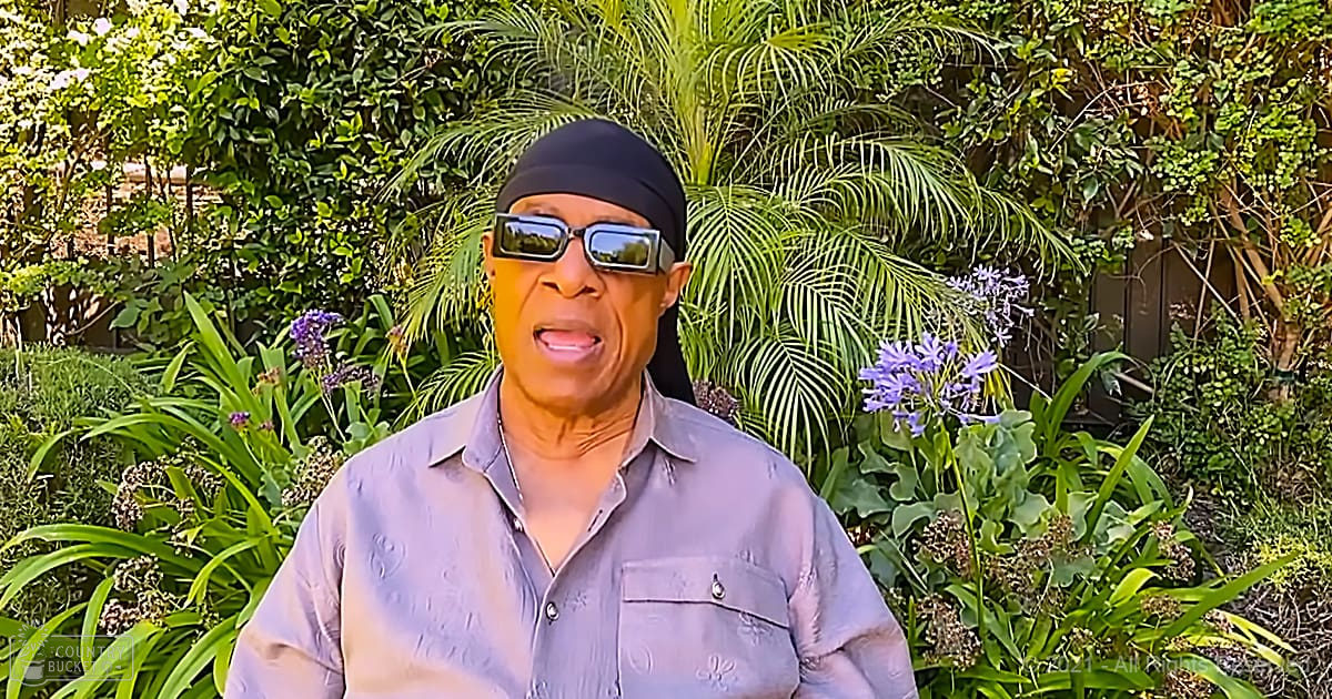 An older adult male stands amidst lush greenery and blooming purple flowers in a garden setting during spring or summer.