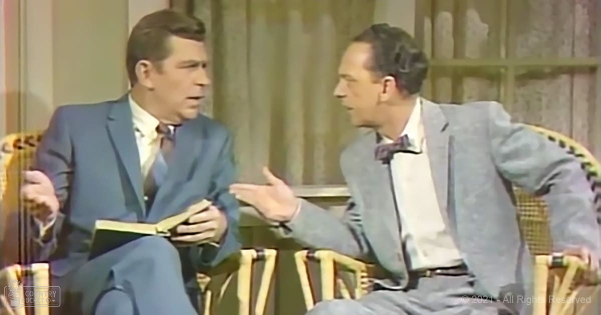 A talk show set with two adult males in formal attire against a light beige curtain and vintage-style wicker chairs.