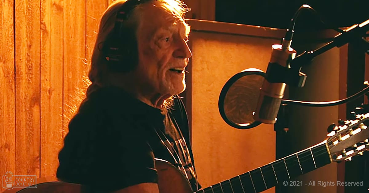 An older adult male with long hair and a beard wearing headphones, a dark t-shirt or sweater in a recording studio environment. The scene is set indoors with wooden paneling on the walls and warm orange lighting.