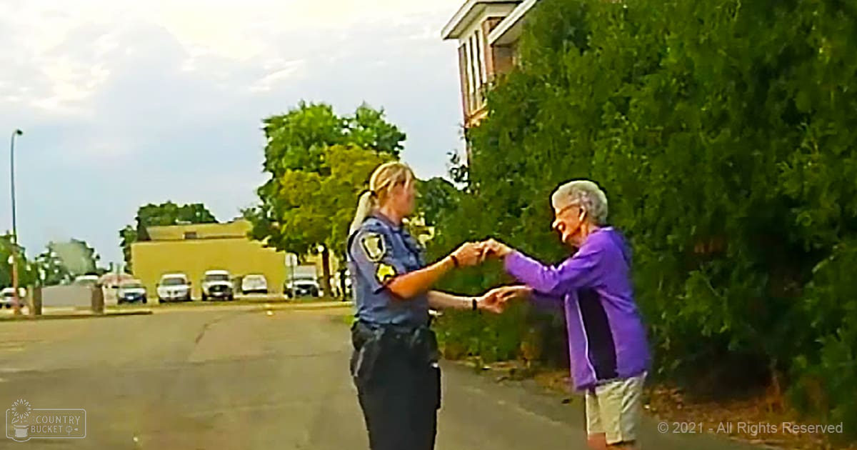 A police officer and an older adult in a suburban neighborhood with lush green trees, yellow and white buildings, and parked cars under bright summer daylight.