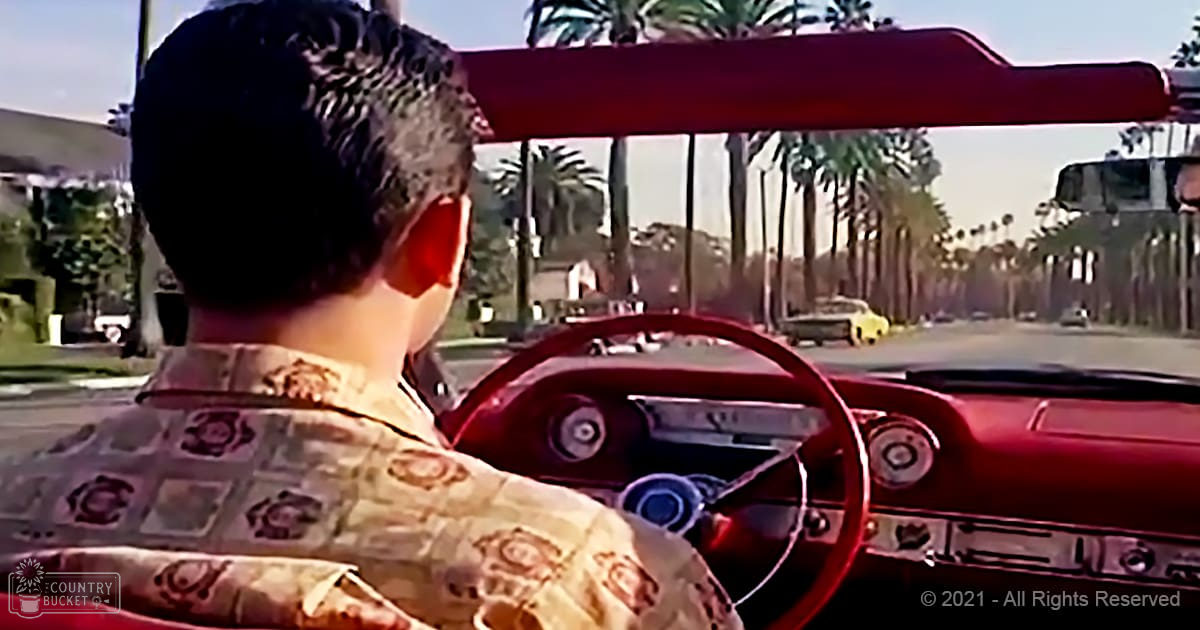 A young adult male drives a classic red car through a suburban neighborhood with palm trees lining the street and houses in the background.