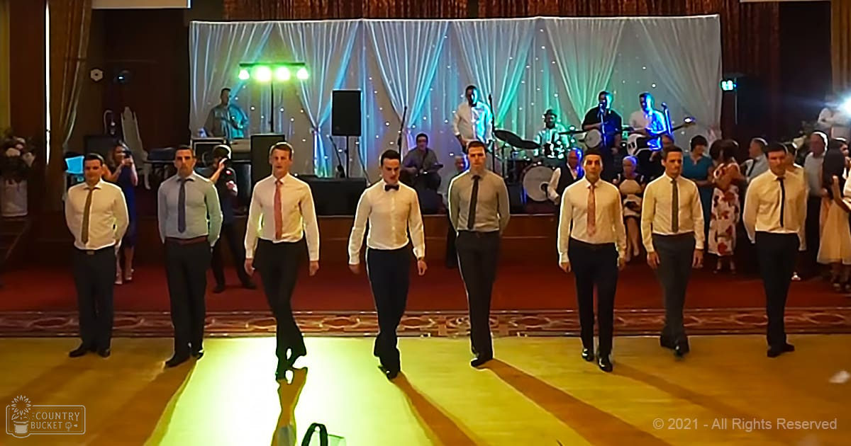 Group of young adult men in formal attire at a wedding reception with musicians on stage, white curtains, and bright lighting.