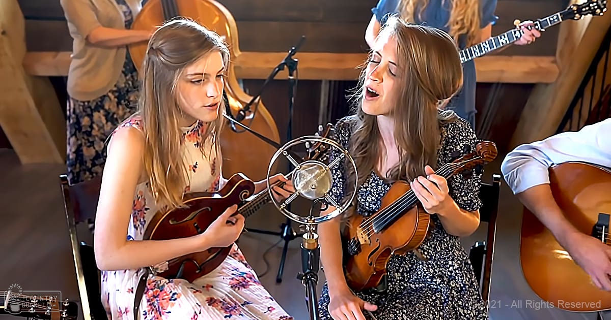Children perform a musical piece indoors, wearing floral dresses; setting appears to be a small studio or performance space.