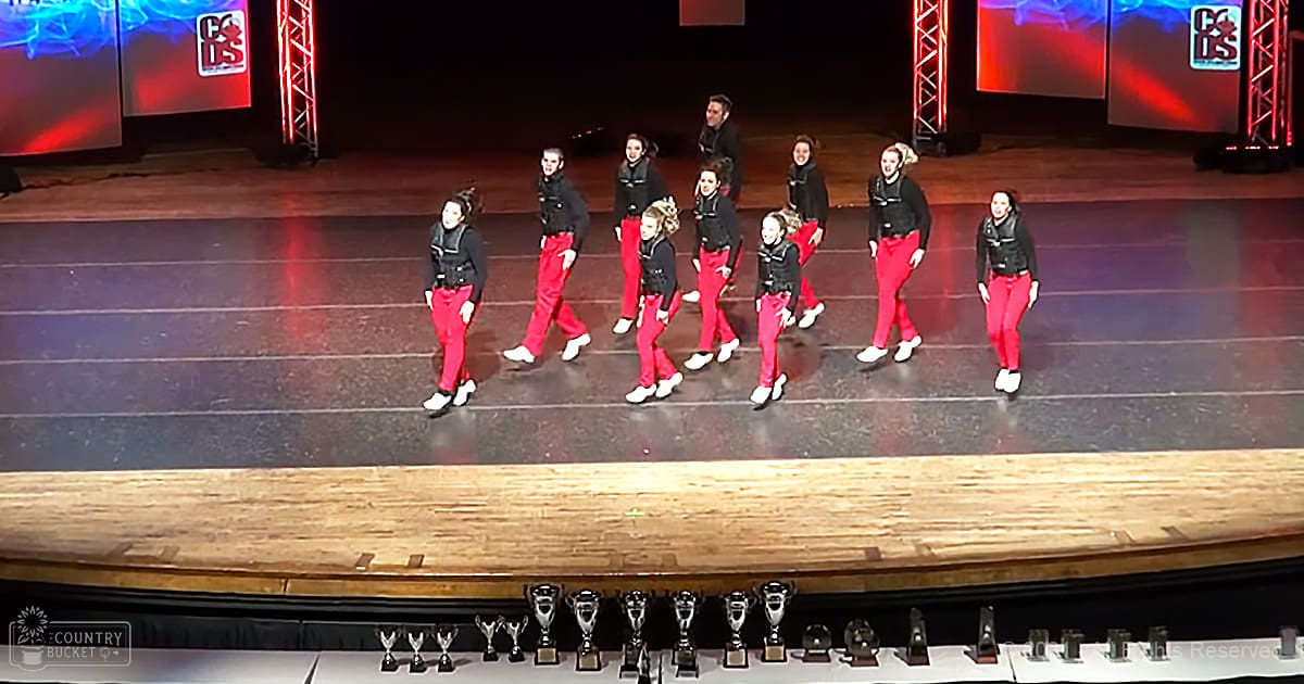 Young adult female dancers in matching black high-collared jackets and red pants performing on a stage with vibrant red and blue lighting effects, indicating an awards ceremony during an evening dance competition.