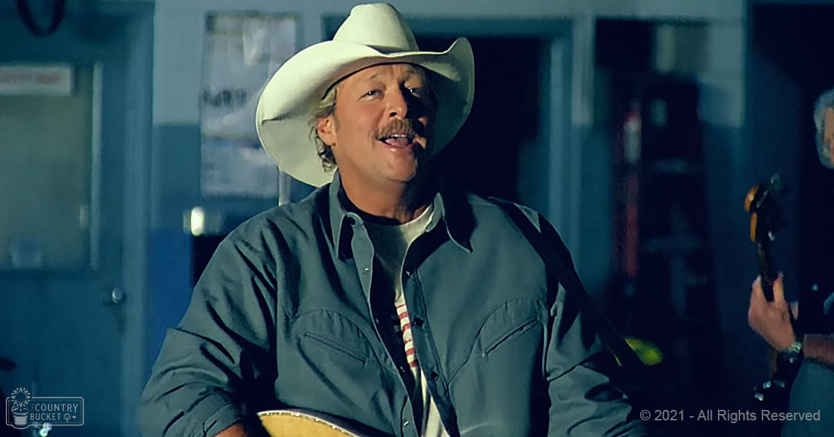 A middle-aged man wearing a light-colored cowboy hat, dark green button-up shirt over striped collared shirt, holding an acoustic guitar in a dimly lit indoor setting with posters and a door frame.