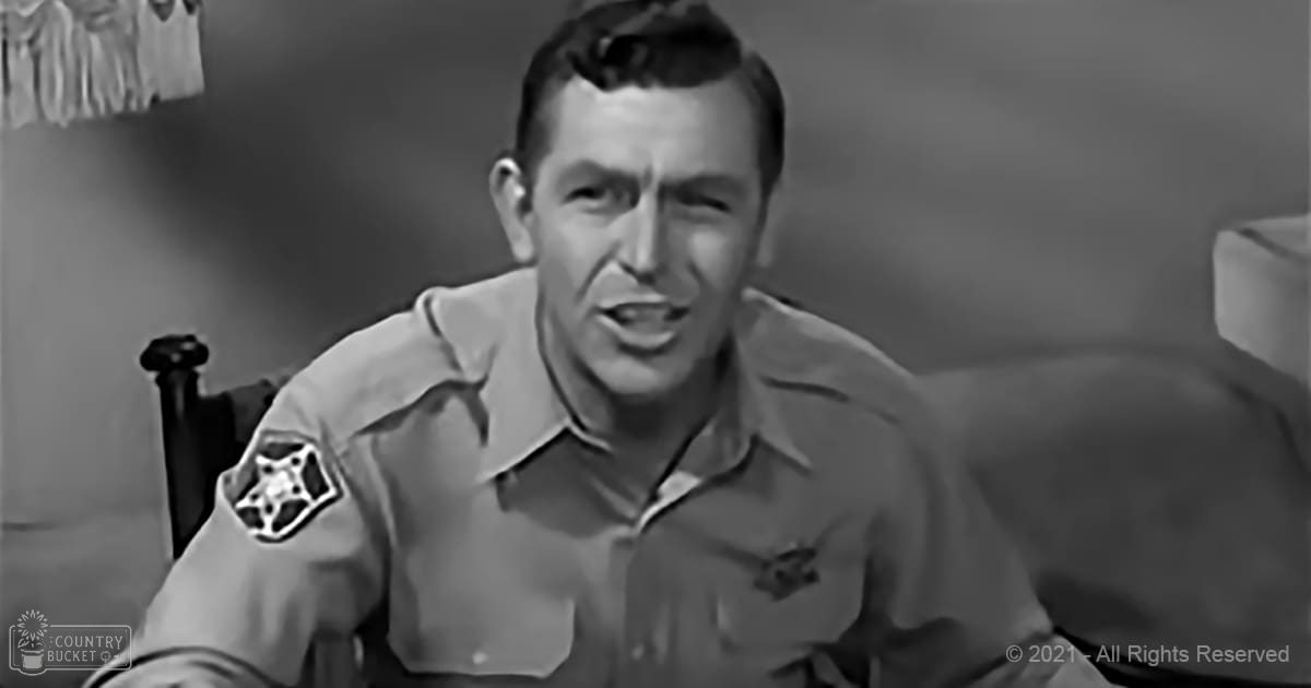 A middle-aged adult male stands indoors wearing a light-colored uniform shirt with patches, suggesting an official or work-related setting. The background includes minimal decor and artificial lighting.