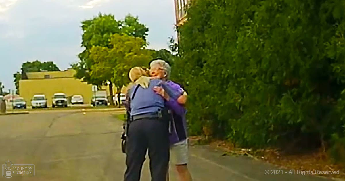 A police officer embracing an older adult amidst a parking lot and yellow building under clear daylight.
