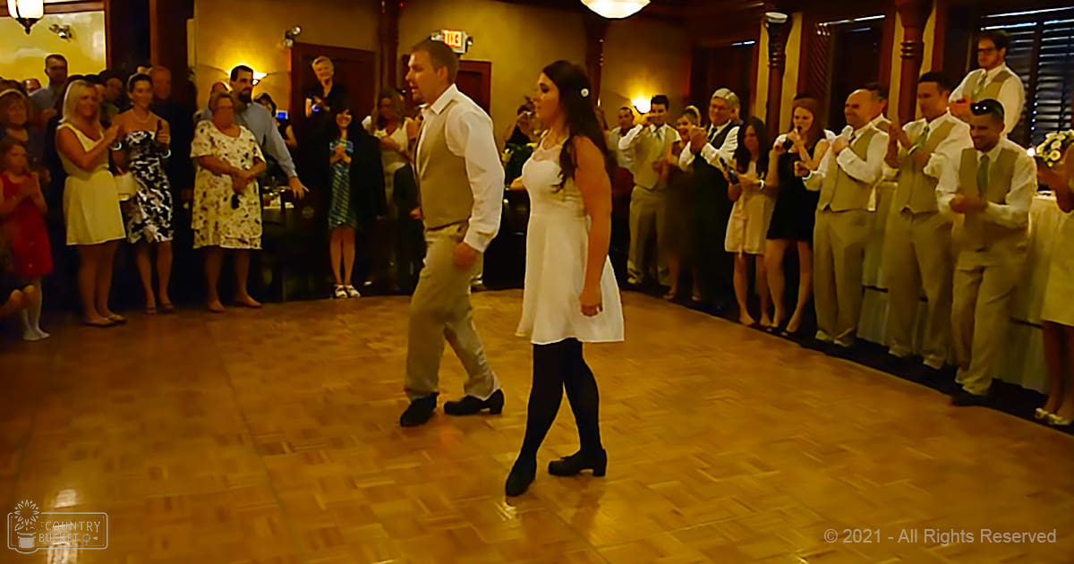 A couple dancing joyfully at what appears to be a wedding reception with an audience clapping and watching in the background, set indoors under warm lighting.