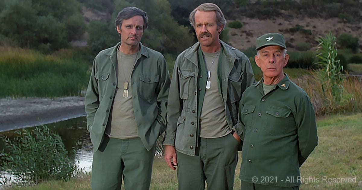 A group of middle-aged men dressed in olive green military uniforms stand outdoors near water, surrounded by grassy areas and trees under natural daylight.