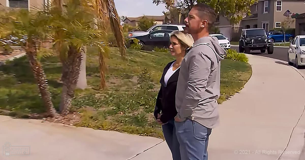 A couple walking in a suburban neighborhood with palm trees lining the sidewalk, an American flag partially visible on one of the houses. The setting suggests spring or summer with greenery and clear skies.