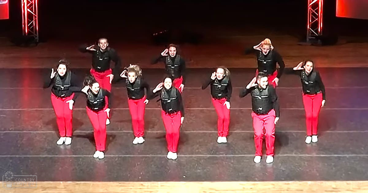 A dance group of young adult females performing on a dark, dimly lit stage with red lighting structures.