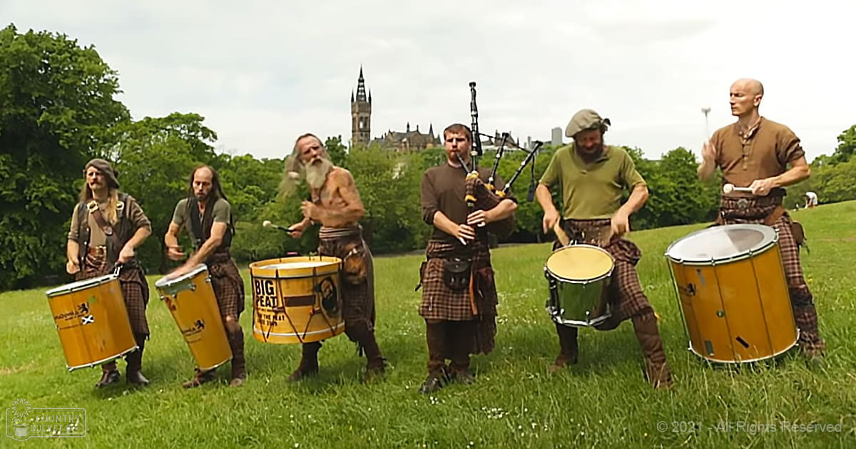 Traditional Scottish music ensemble performing outdoors in a park-like setting with a castle in the background, with bagpipes and drums under an overcast sky.
