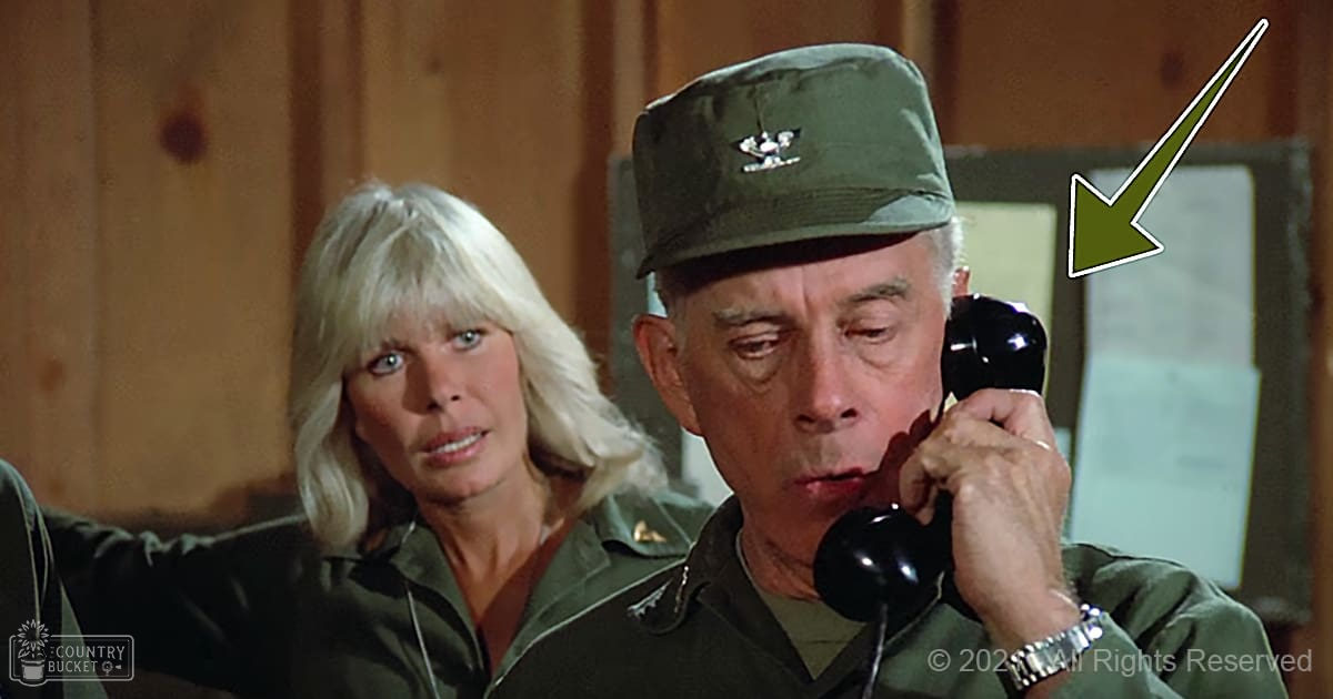 A man wearing a military uniform, holding an old-fashioned telephone indoors with wooden paneling, suggesting a mid-20th-century film setting.