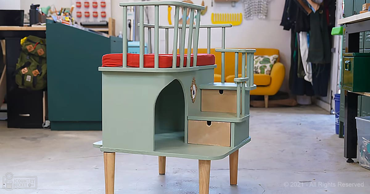 A repurposed chair with a greenish-blue paint job, red cushion, and beige drawers serves as a pet house in an indoor workshop or garage setting with various tools and storage items visible.
