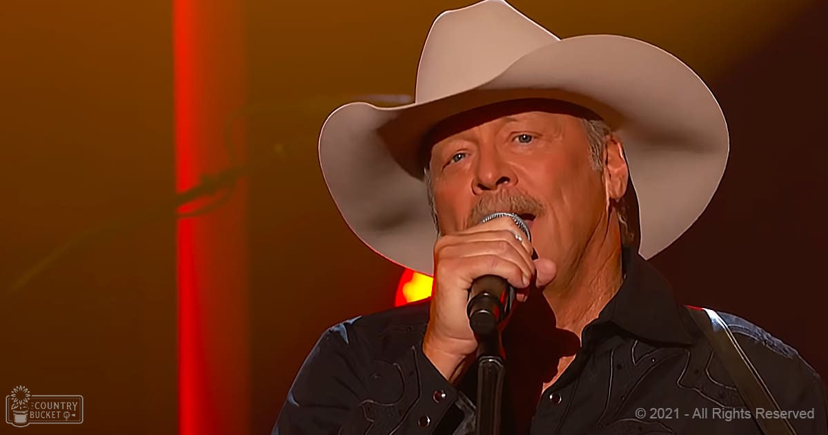 A middle-aged adult male singer wearing a black button-up shirt with decorative stitching and a white cowboy hat, performing on stage under warm red lighting at an indoor concert venue.
