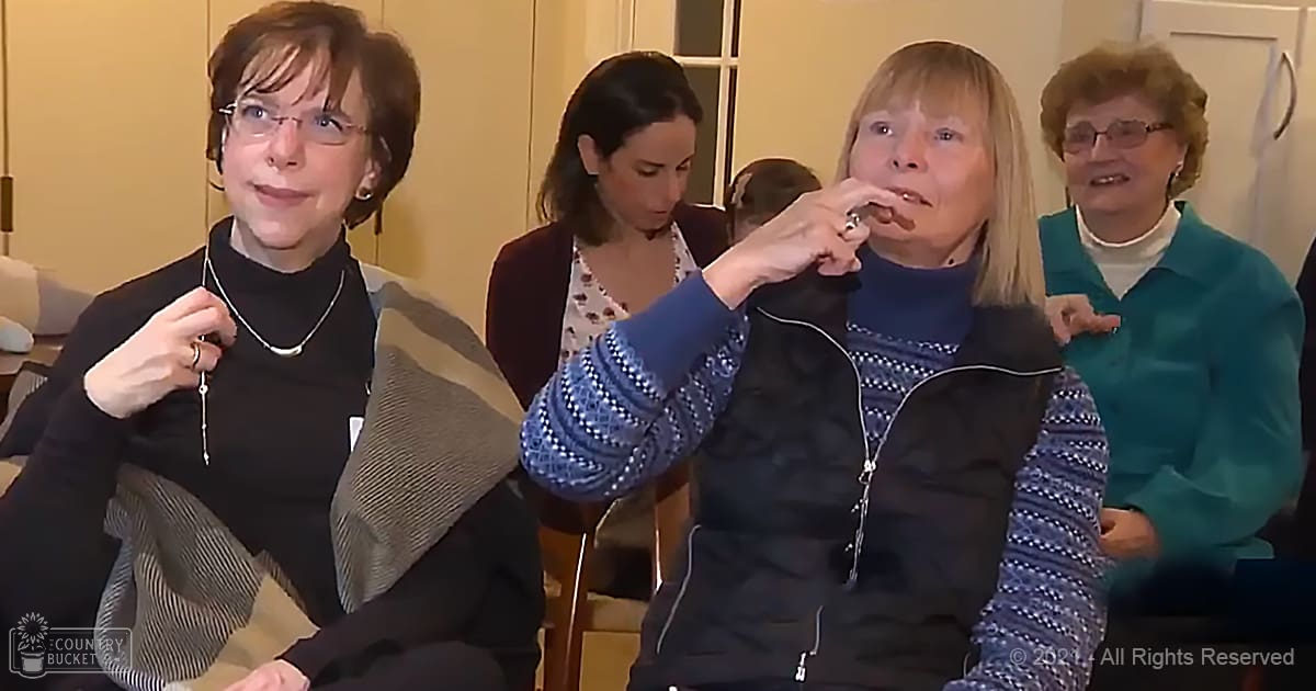 A middle-aged woman in a living room setting, wearing a black turtleneck sweater with a blue and white zip-up jacket over it, accessorized with a silver necklace.