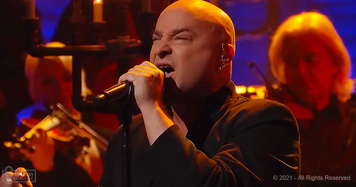 An adult male singer in a dark V-neck shirt performing on stage with other musicians playing string instruments under dim blue lighting.