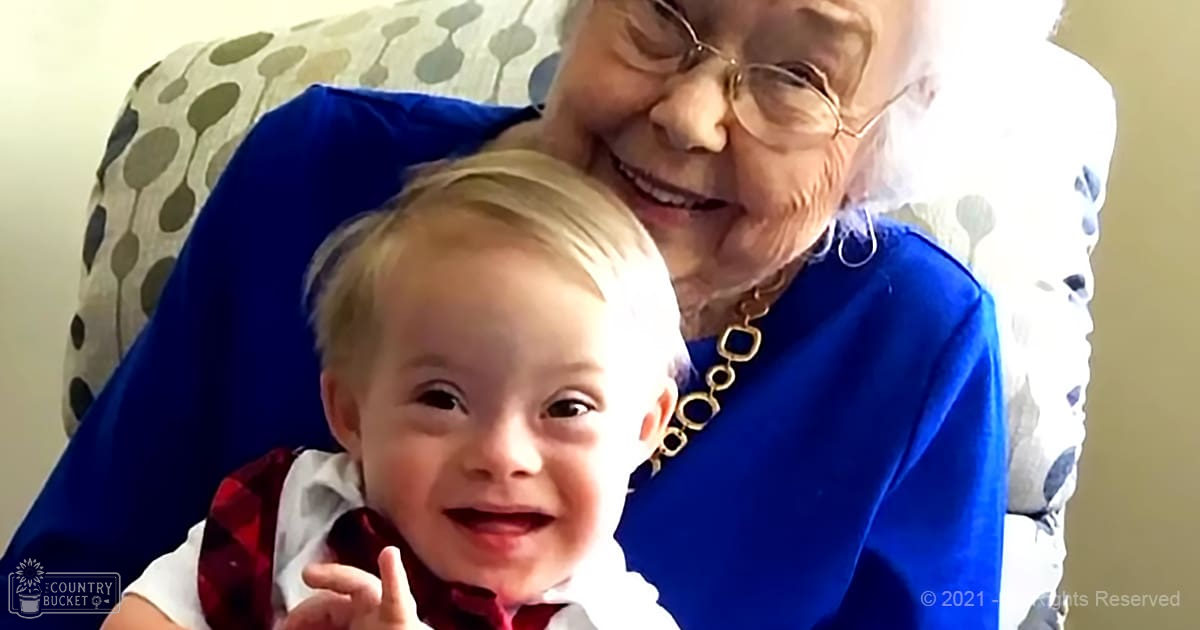 A toddler in a white shirt with red patterned vest sits indoors near an older adult wearing a blue dress and gold necklace, surrounded by furniture like a patterned chair or couch.