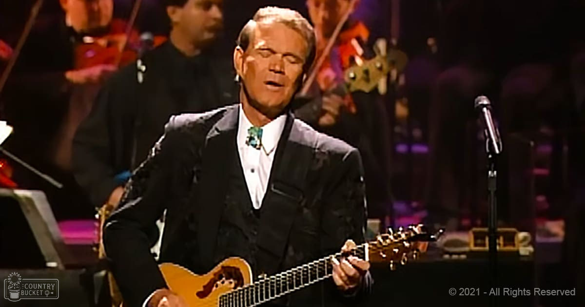 Middle-aged man in dark suit jacket and white shirt performing on stage at an indoor concert venue, surrounded by musical instruments and sheet music.