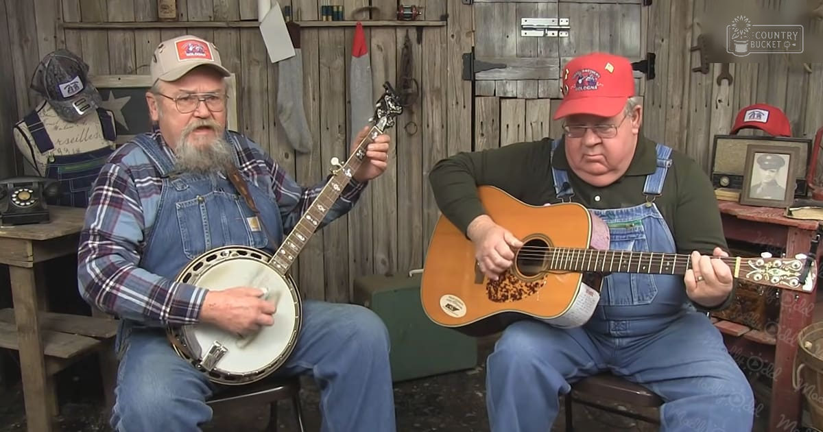 Two older adult men playing musical instruments in a rustic indoor setting with wooden furniture, decorative objects, and natural daylight.