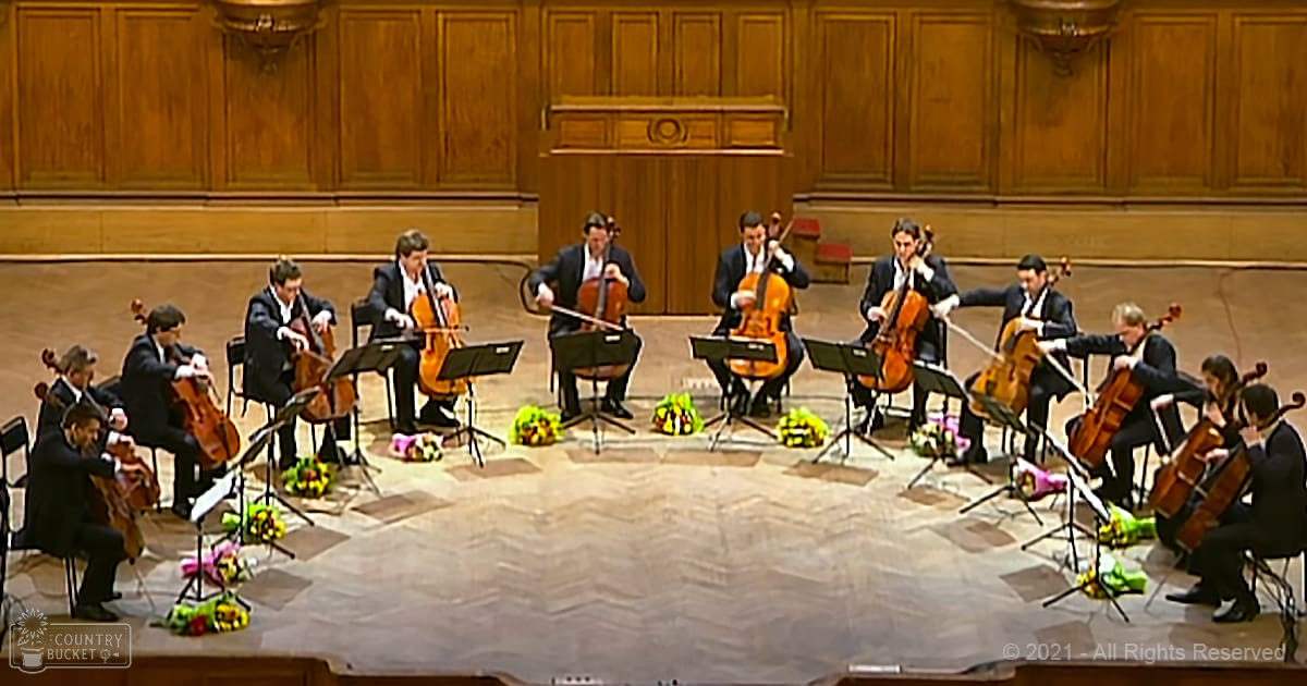 A string quartet performs in a brightly lit indoor concert hall with polished wooden floors, surrounded by musicians wearing dark suits and white shirts. Wooden paneling on walls and small bouquets of flowers adorn each chair.