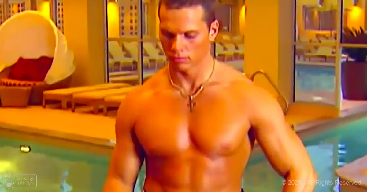 A muscular adult male with a necklace stands shirtless by an indoor pool area, surrounded by lounge chairs and plants.
