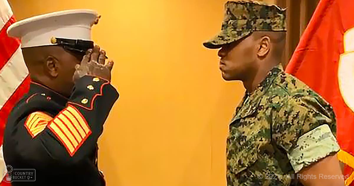 An adult male singer wearing a dark blue dress uniform with gold stripes and insignia, standing alongside another adult male in a camouflage-patterned military uniform during what appears to be an indoor military ceremony.