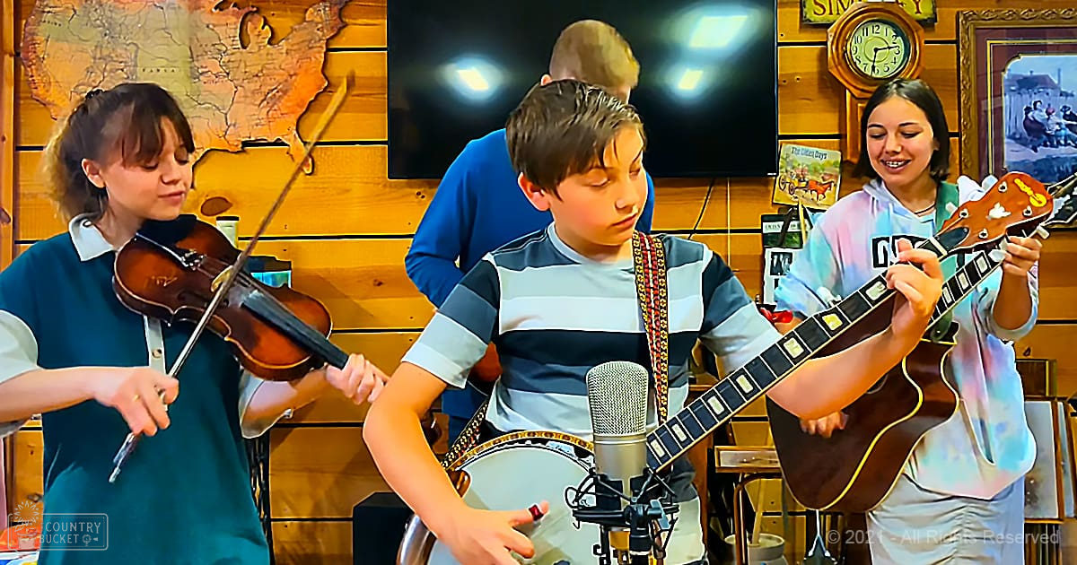 Three children playing music in a home studio with teal dresses, striped shirts, and tie-dye hoodies, surrounded by musical instruments and indoor decor.