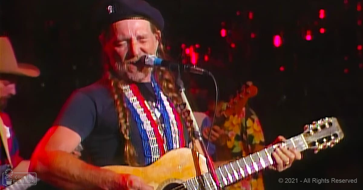 A middle-aged male performer wearing a patterned red, white, and blue scarf around his neck, a black cap, and possibly a wristwatch or bracelet on the left wrist, stands in an indoor concert setting with stage lights visible behind him.