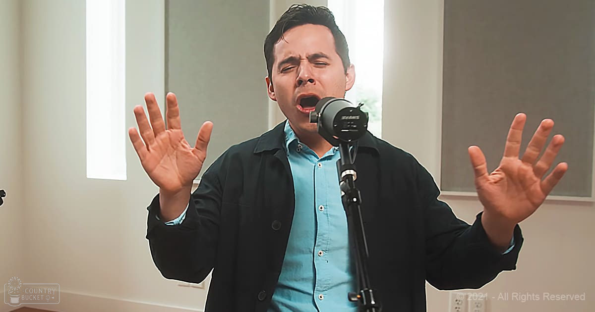 A young adult male singing in a modern indoor recording studio with natural light streaming through the window, neutral walls, and professional equipment.