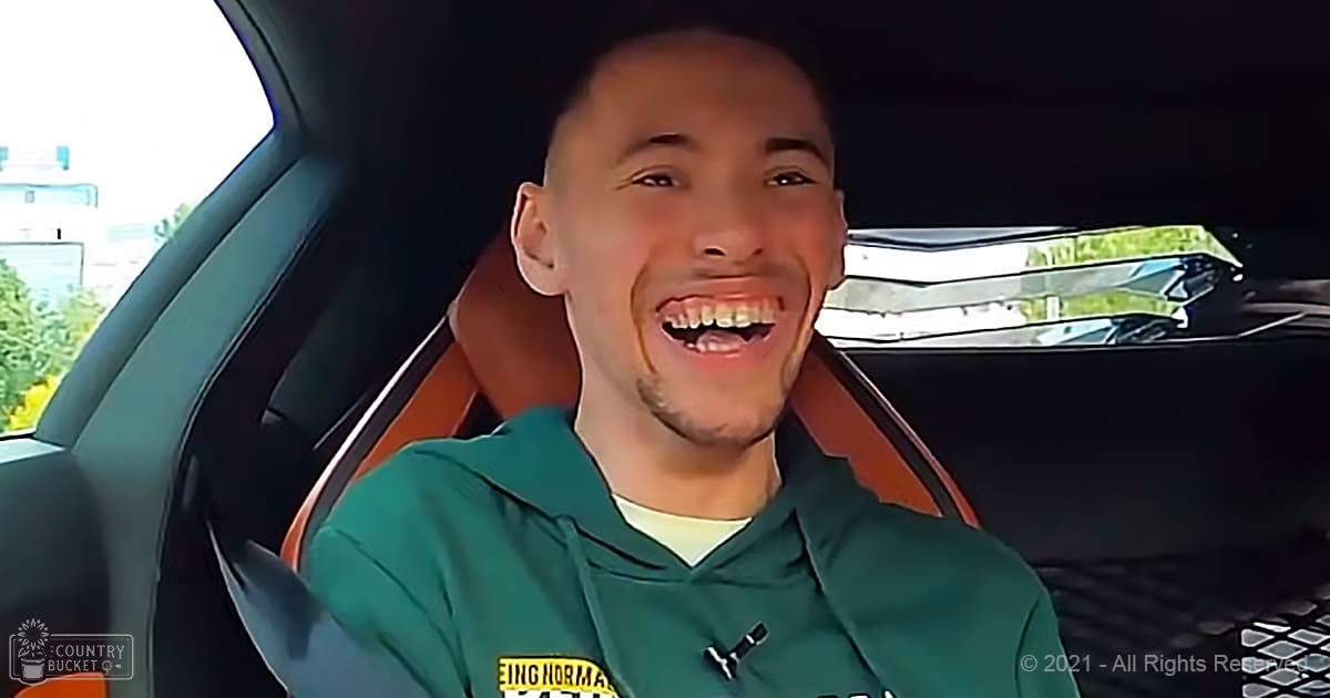 A young adult male in a relaxed fit green hoodie with yellow and black text, sitting inside an orange-seat car at what appears to be a parking lot or roadside.