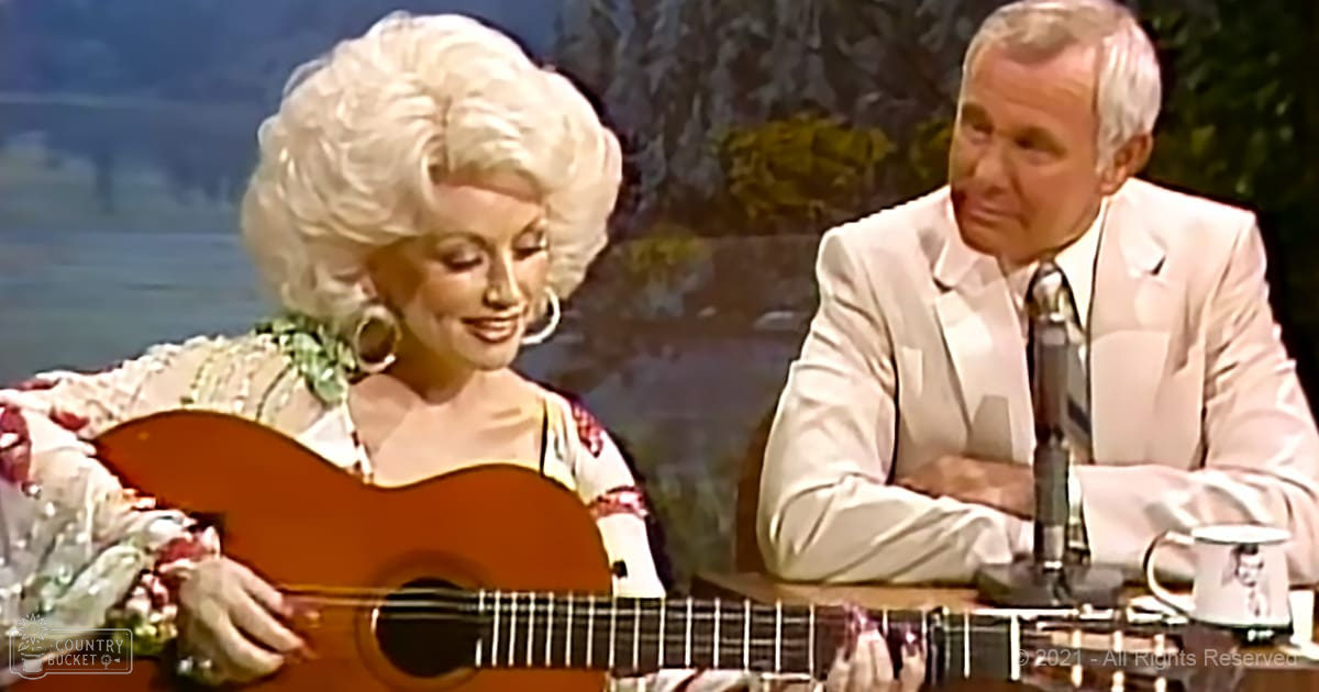 A woman playing a natural wood acoustic guitar with an older adult man seated next to her in a studio setting, wearing a floral-patterned top or dress and large hoop earrings; the backdrop suggests spring or summer.
