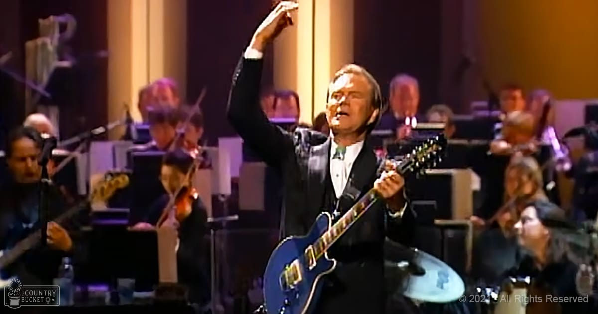 A middle-aged man playing a blue electric guitar in front of an orchestra within a classical concert hall under warm artificial lighting.