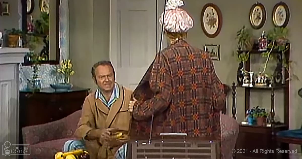 Middle-aged man wearing a brown jacket with striped shirt and light pants sits indoors holding an object, older woman in plaid coat stands nearby with decorative hat, vintage-style furniture and decor fill the room, natural daylight illuminates the space.