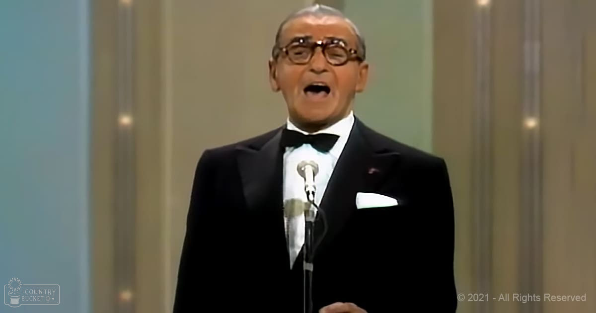 A middle-aged adult male in a black tuxedo with a white shirt and bow tie, set against an indoor stage backdrop with vertical light panels.