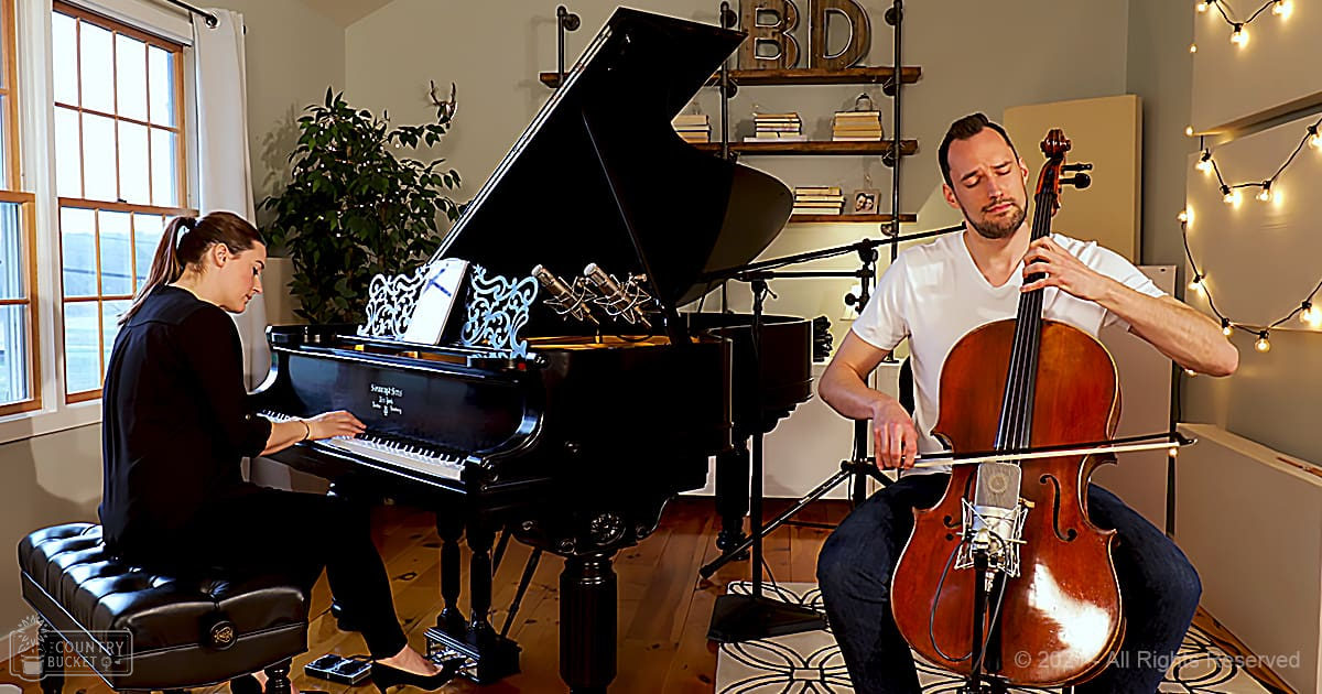 Musical performance scene in an indoor music studio with a young adult female singer wearing a black dress and wristwatch, alongside an adult male musician in a white t-shirt. Grand piano and string lights create a cozy ambiance.