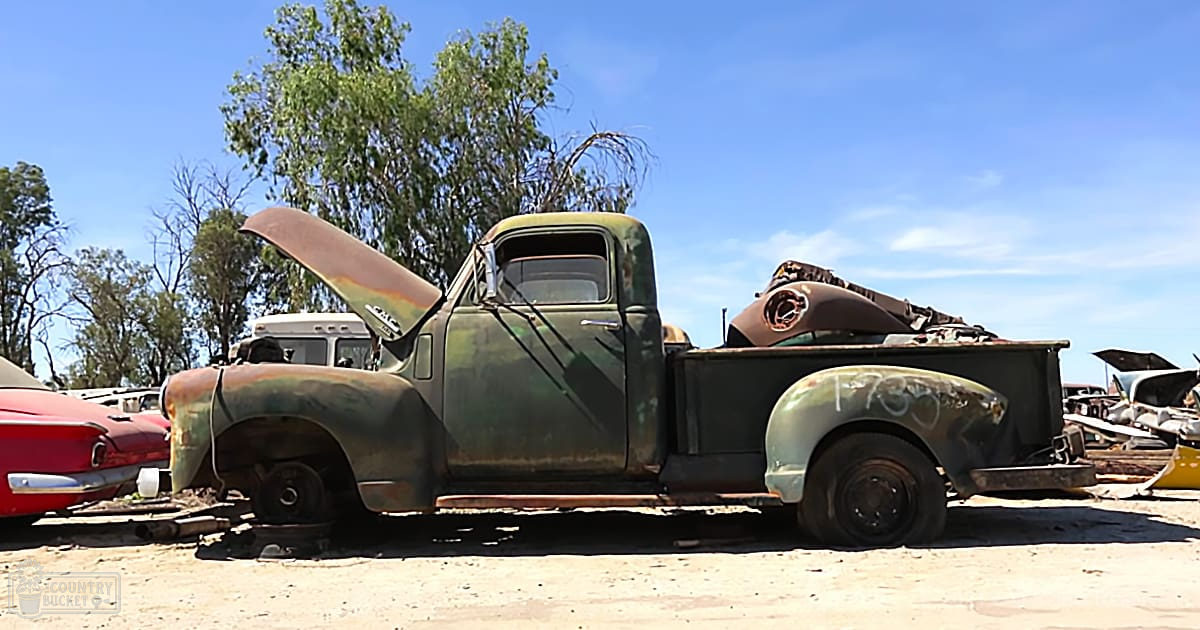 An old, rusted pickup truck in a junkyard with trees and a clear blue sky indicating late spring or summer daylight hours.