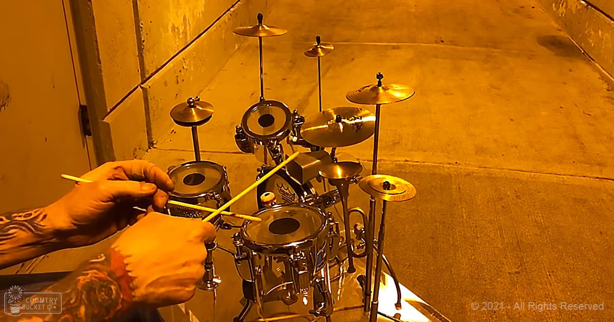 A young adult male drummer with tattooed forearms plays an electronic drum set in a basement or garage setting under yellowish-orange lighting.