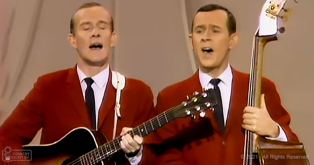 An adult male singer in a red suit jacket with white shirt and dark tie, holding an acoustic guitar. Another adult male performer to his right also wearing a similar outfit and holding a double bass against a plain light-colored background.
