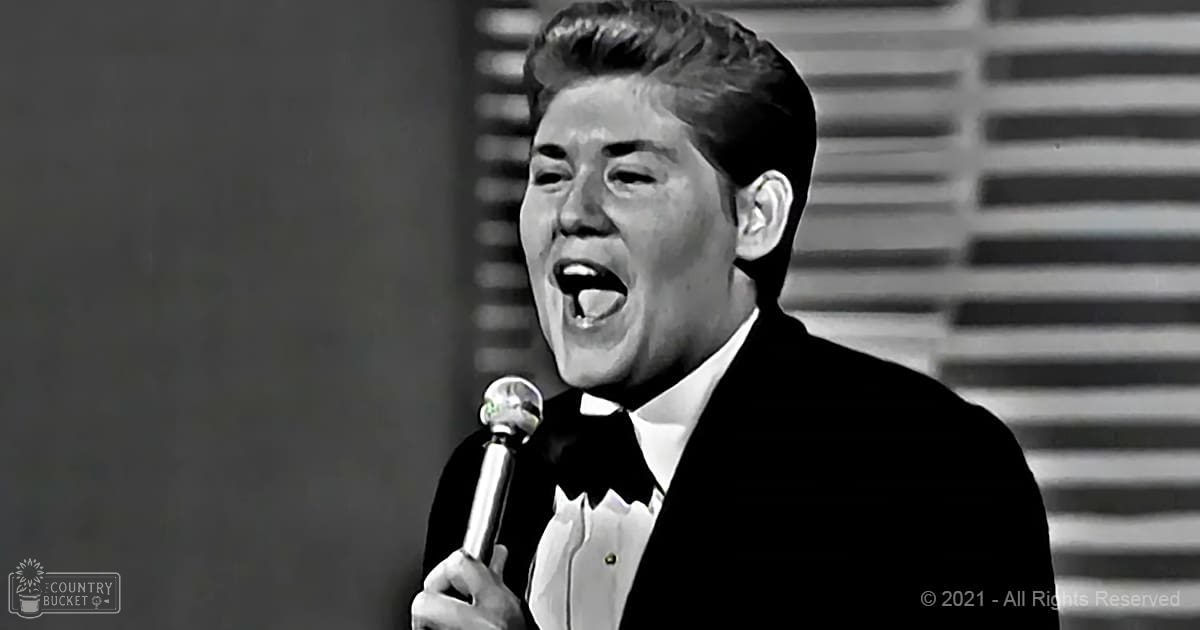 Adult male singer in a black suit jacket and white shirt with a bow tie, holding a microphone in a studio setting.