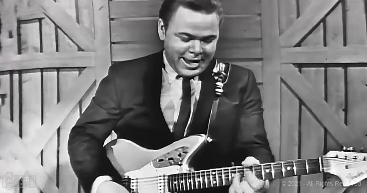 A young adult male playing guitar in an indoor setting with wooden paneling, wearing a dark-colored suit and a patterned tie. The image is black-and-white.