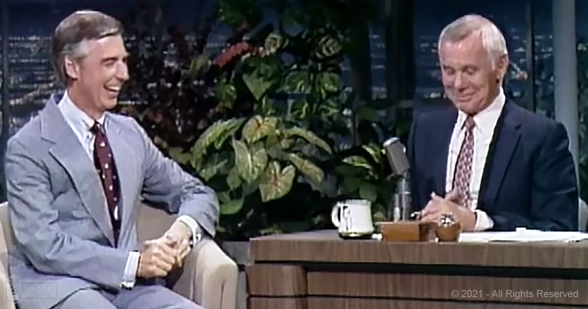 Talk show set in an indoor studio with two older adult males wearing mid-20th century attire; wooden desk with microphone, mug, and papers; decorative plants behind subjects.