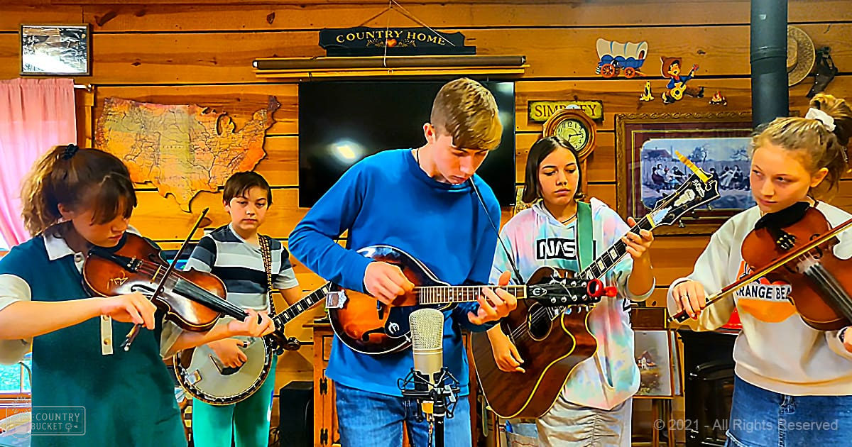 A group of young musicians performing in a country home setting with wooden walls, a map of the United States, and natural light streaming through.