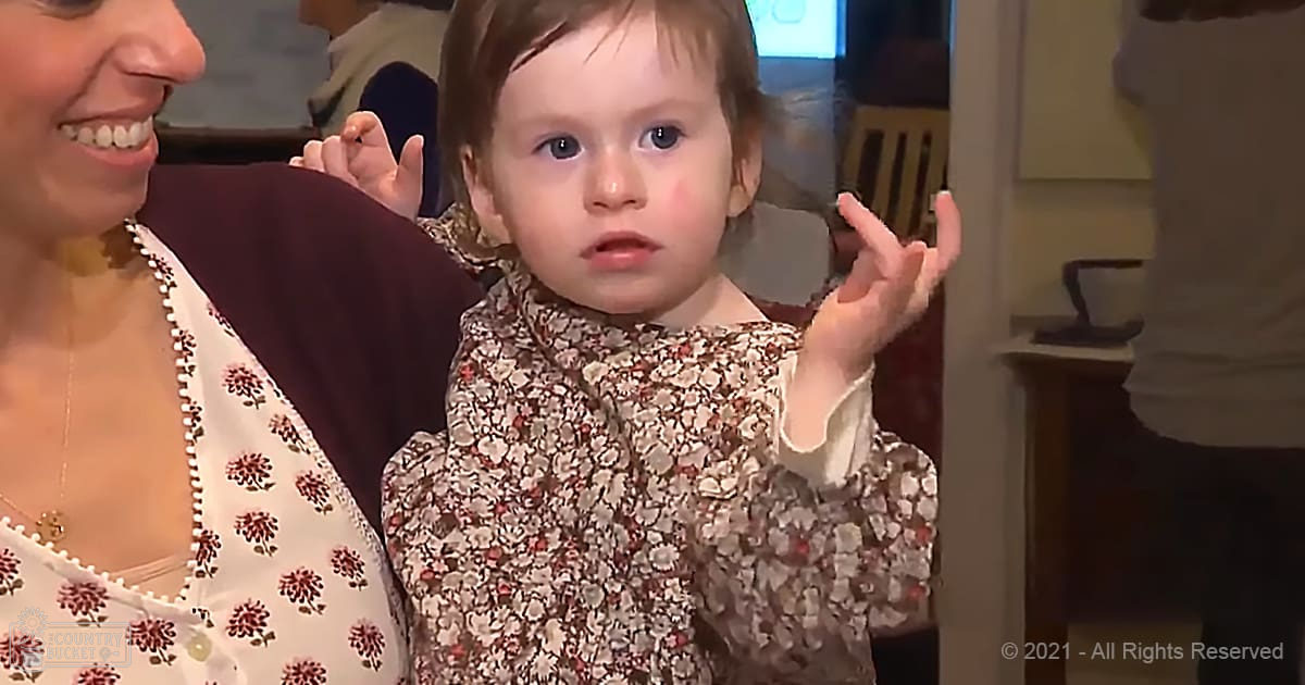 A toddler wearing a floral dress with brown, white, and red patterns stands indoors next to an adult woman in a floral top; the scene is softly lit.