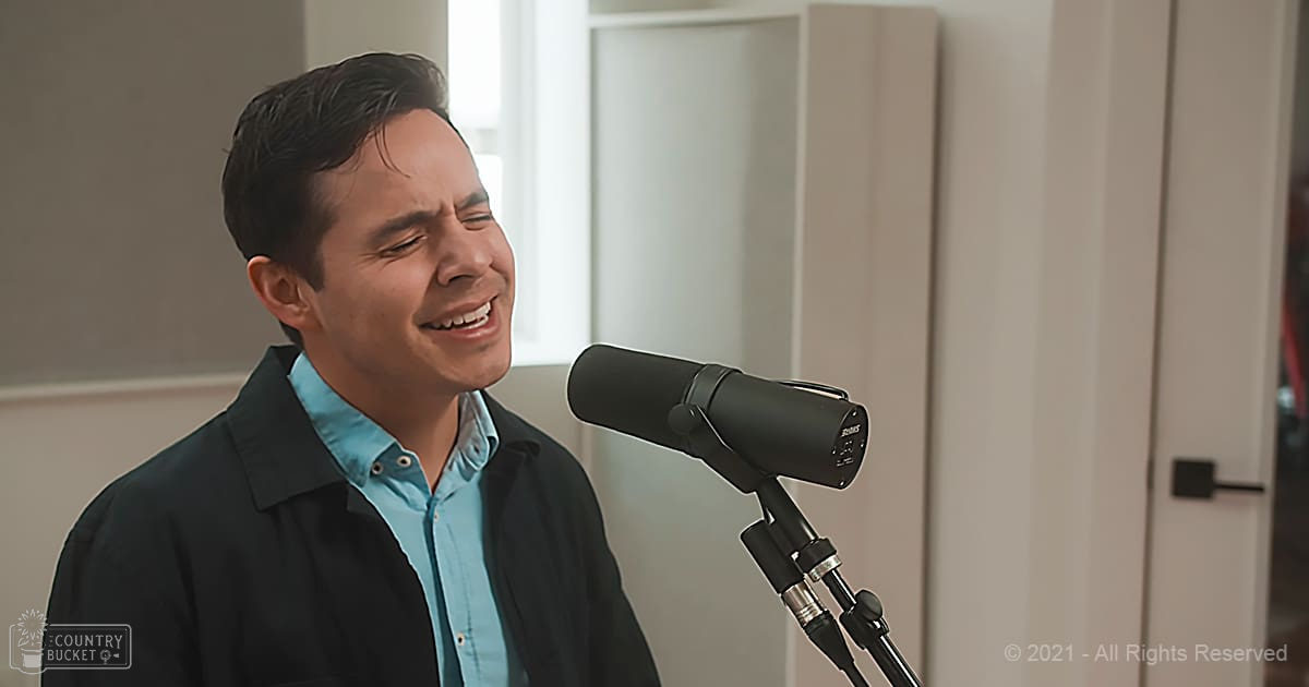 Young adult male singing into a microphone in an indoor recording studio with black jacket, light blue button-up shirt, and natural light coming through the window.