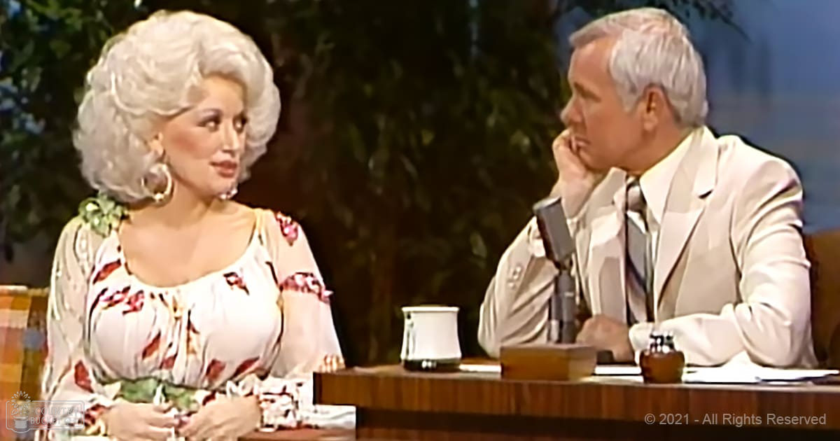 An adult female in a floral-patterned blouse with an older adult male in a white suit jacket seated at a wooden table during what appears to be a mid-20th-century television interview setup.