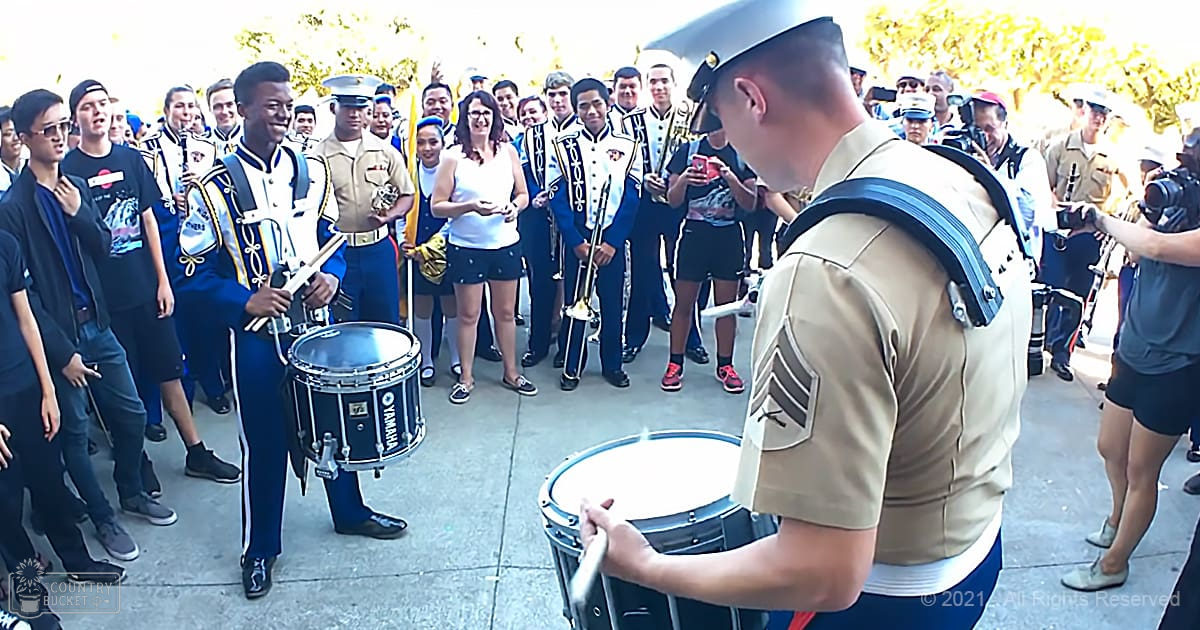 Young adults and middle-aged individuals in a marching band uniform performing outdoors, with focus on drummers and civilian attendees recording or photographing the event.