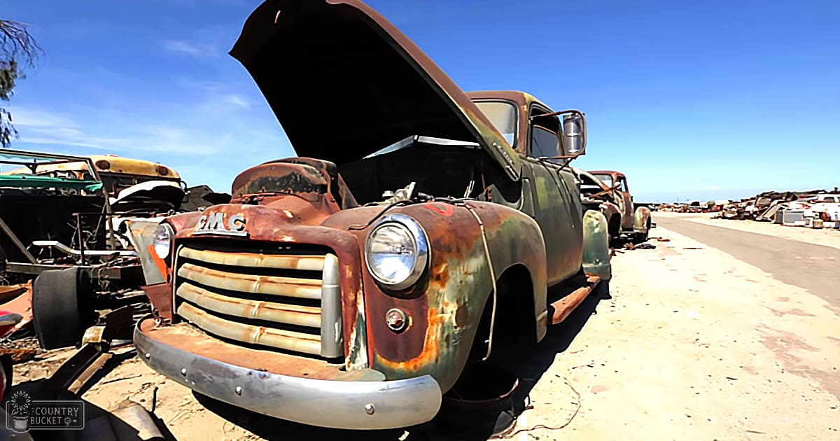 Rusty old GMC truck in junkyard with bright sunlight, outdoor setting.