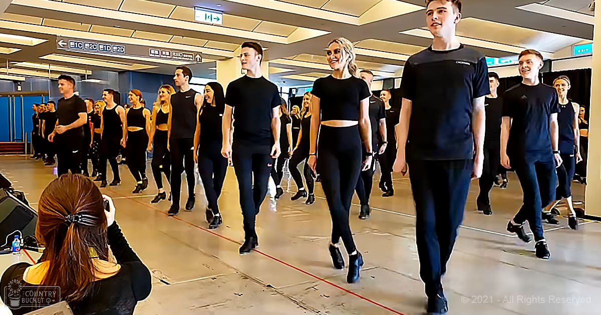 A dance class in progress at an indoor studio with mirrors, signs indicating room numbers, and a polished floor marked by red lines.