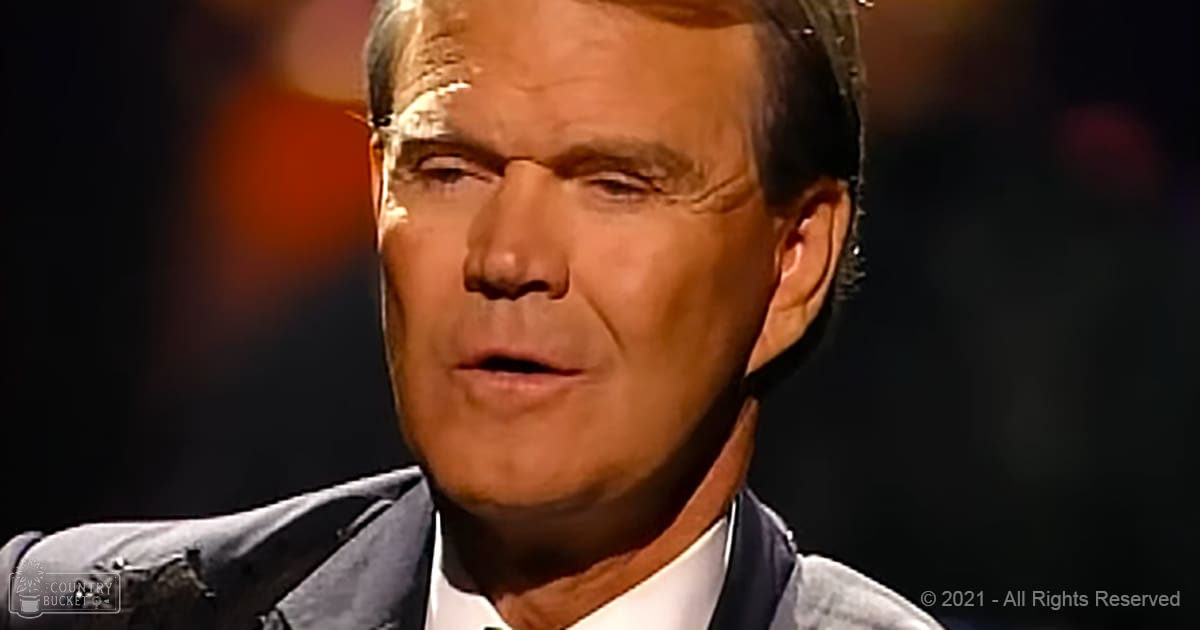 Middle-aged adult male wearing a dark gray or navy blue tailored suit jacket with lapel pin, white collared shirt, and formal attire, set against an indoor stage environment with professional lighting.